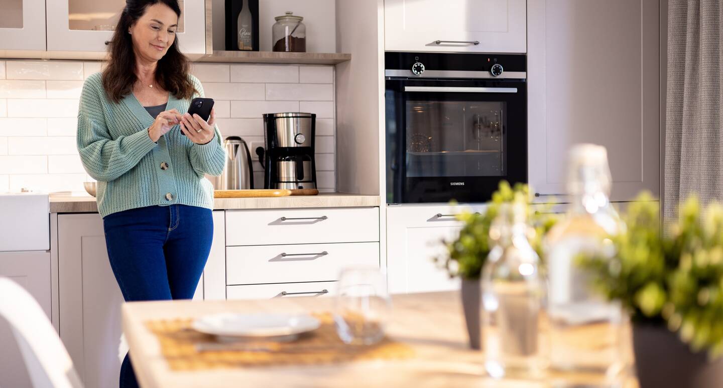Eine Frau steht in ihrer Küche und schaut lächelnd auf ihr Smartphone