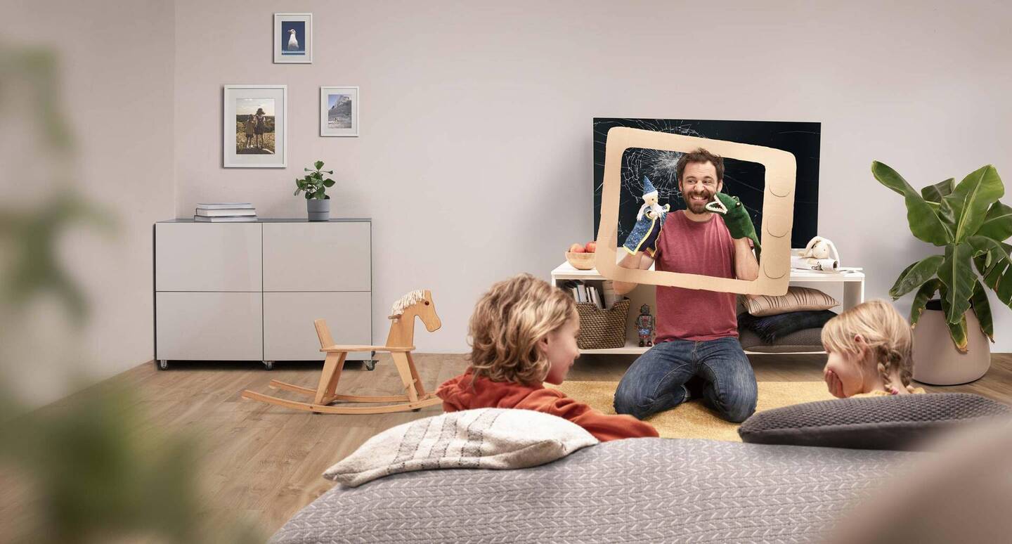 Eine Wohnzimmersituation, in der ein Vater ein Puppentheater vor einem kaputten Fernseher spielt