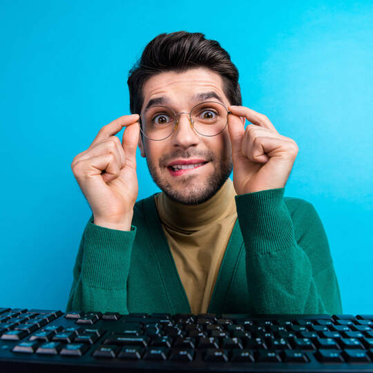 Ein Mann sitzt vor einer Tastatur und fasst sich an seine Brille. 